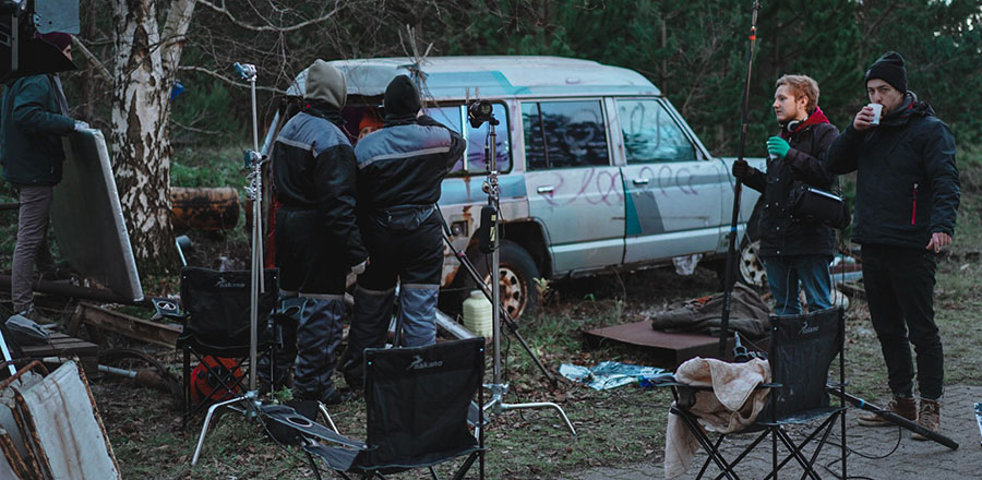 En gruppe unge mænd står udendørs i varme heldragter på et filmset, omgivet af udstyr.