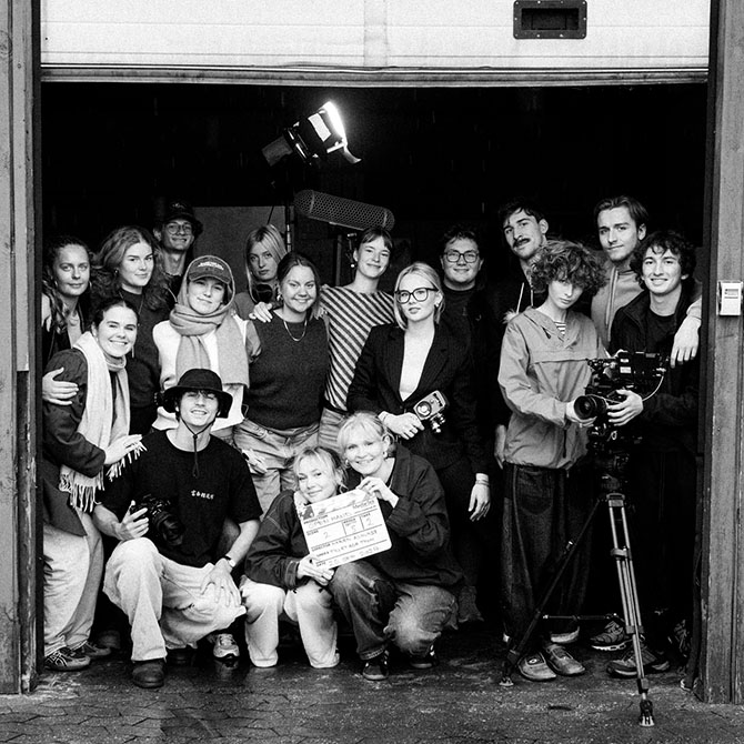 A group of young people are standing close together at the entrance to a storage building. They have a clapboard and camera and are all smiling.